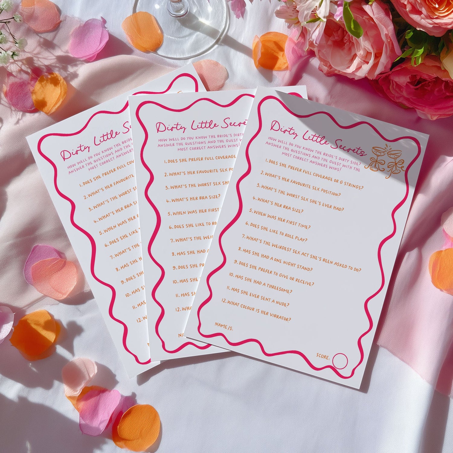 Three white wedding guest books with pink wavy borders and gold script titles are arranged on a white surface with scattered rose petals and pink flowers. The guest books have pink and gold printed text and check boxes.