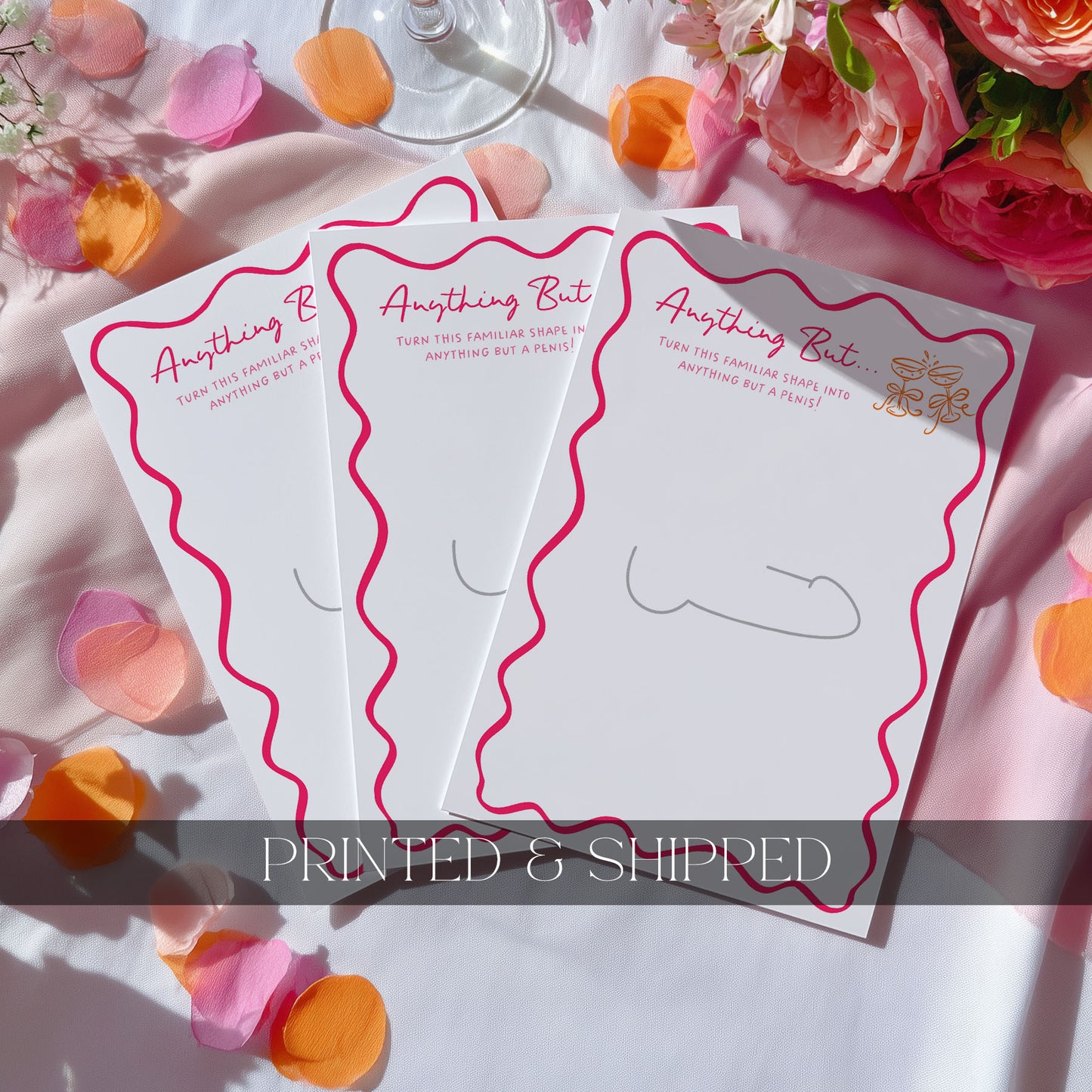 Four Anything but drawing game cards with pink wavy borders and gold foil accents, arranged on a white fabric background with pink and orange flower petals and roses.