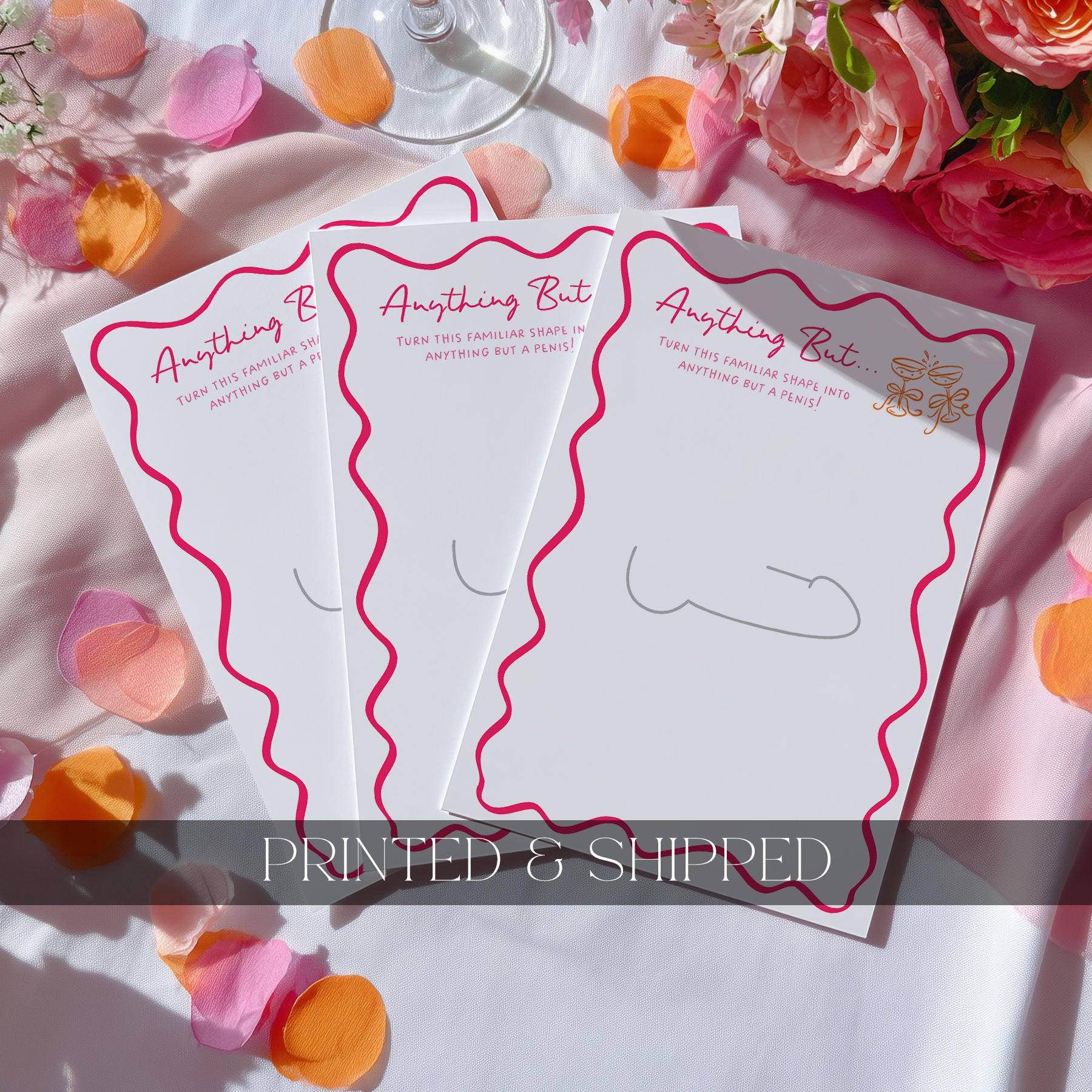 Four Anything but drawing game cards with pink wavy borders and gold foil accents, arranged on a white fabric background with pink and orange flower petals and roses.