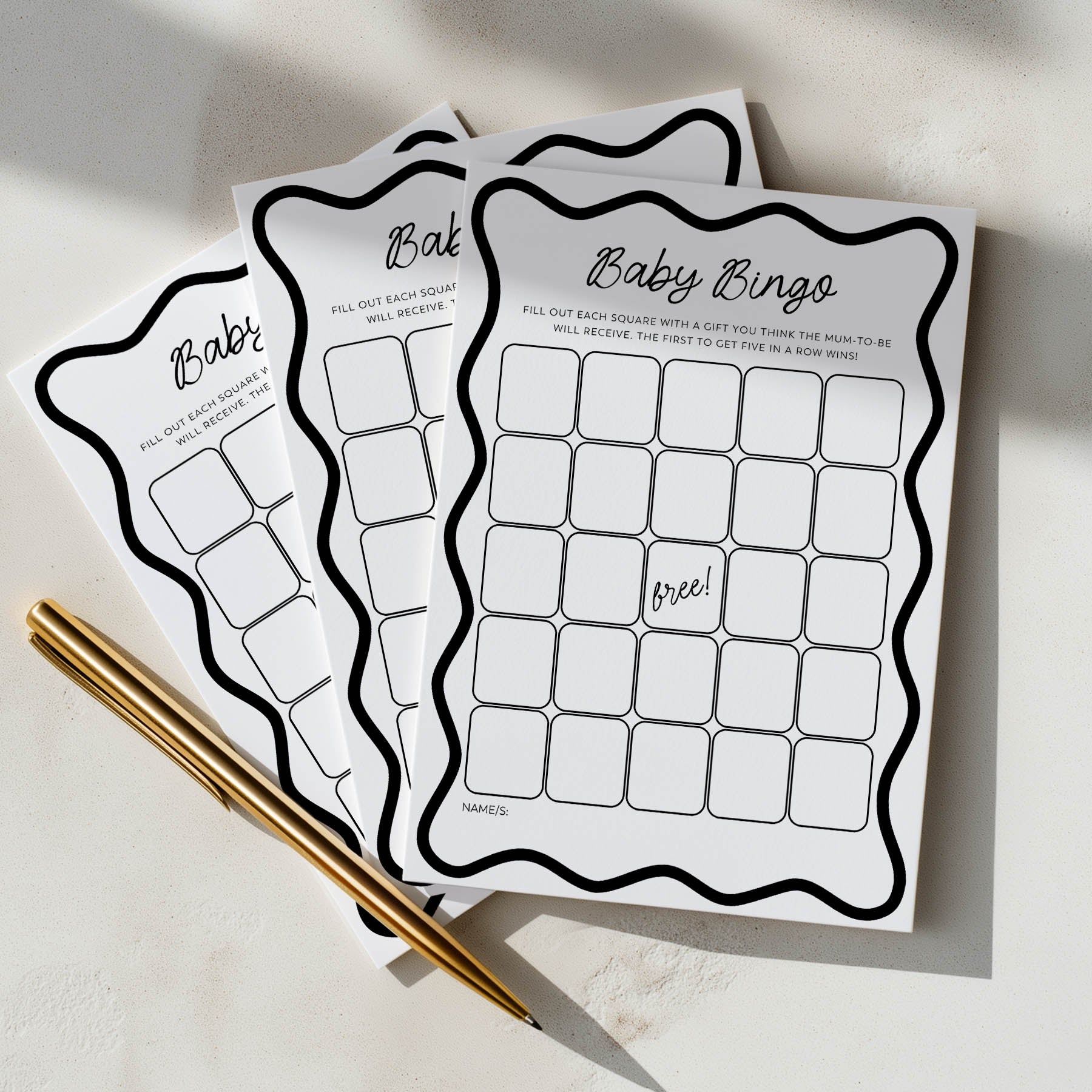 Three baby bingo cards on a light surface with a gold pen.