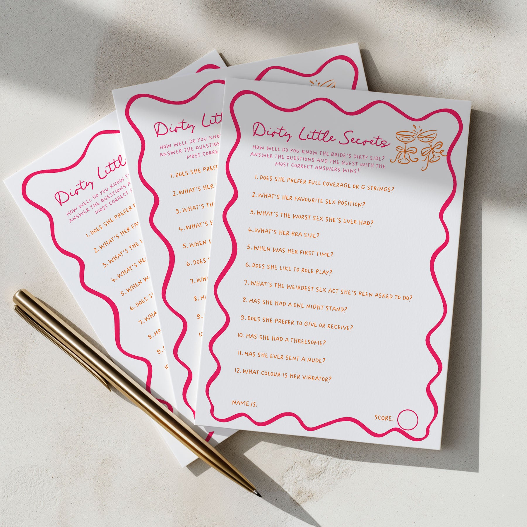 Three white party game cards with pink wavy borders and gold text reading 'Dirty Little Secrets' are arranged on a light surface with two gold pens.