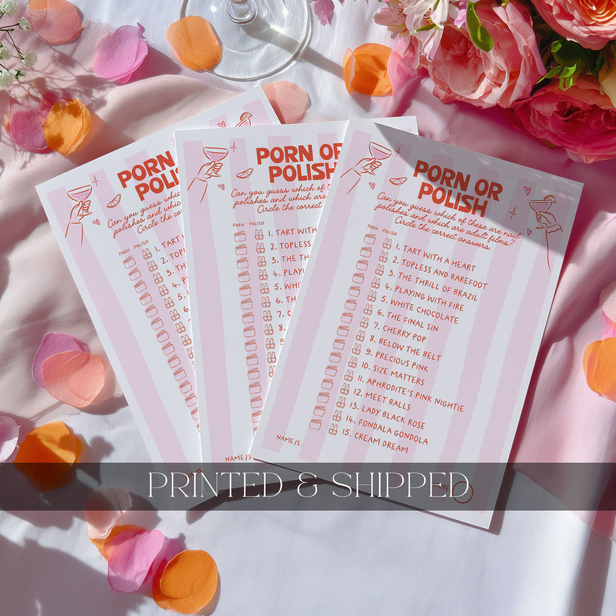 Three pink and white 'Would She Rather?' party games cards are displayed in a flat-lay arrangement with rose petals, flowers, and a wine glass on a white tablecloth. A dark gray banner at the bottom reads 'PRINTED & SHIPPED'.