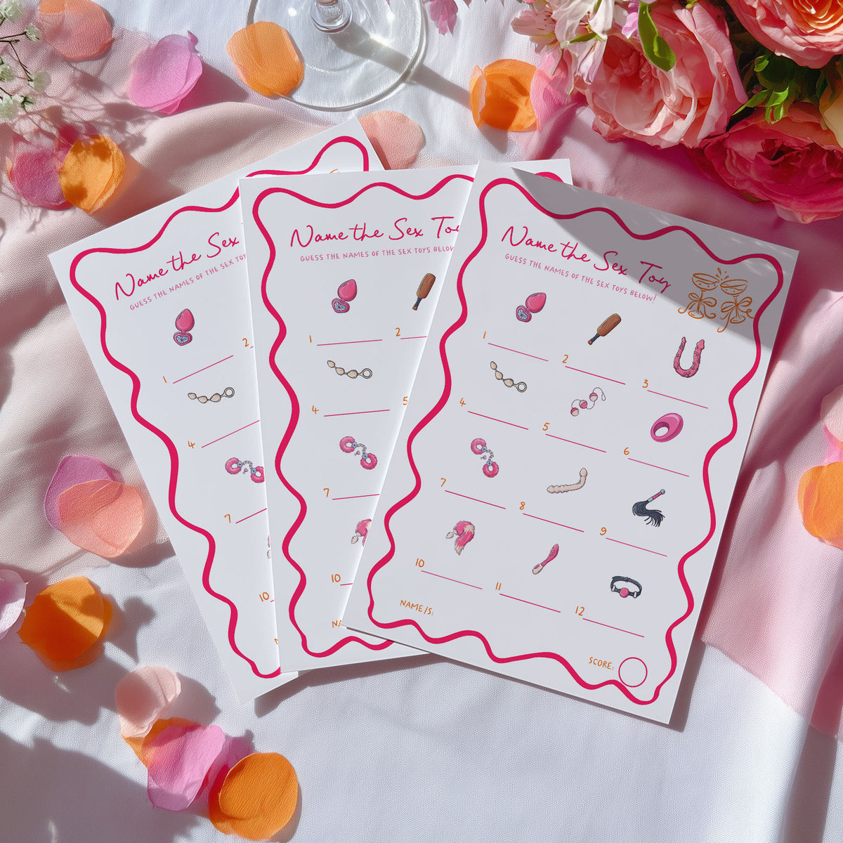 Three white game cards with pink wavy borders and pink-themed clues are arranged on a white fabric surface with pink and orange flower petals and flowers around them.