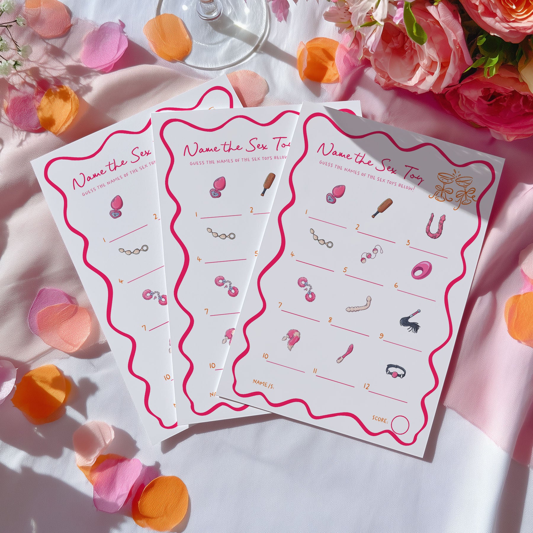 Three white game cards with pink wavy borders and pink-themed clues are arranged on a white fabric surface with pink and orange flower petals and flowers around them.