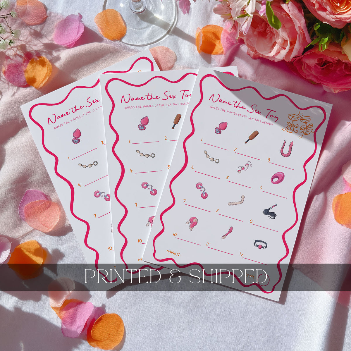 Three printed game cards titled 'Name the Sex Toy' are arranged in a fan shape on a white satin fabric with pink rose petals and orange flower petals around them. The cards feature pink wavy borders and small illustrations of intimate accessories with numbered prompts.