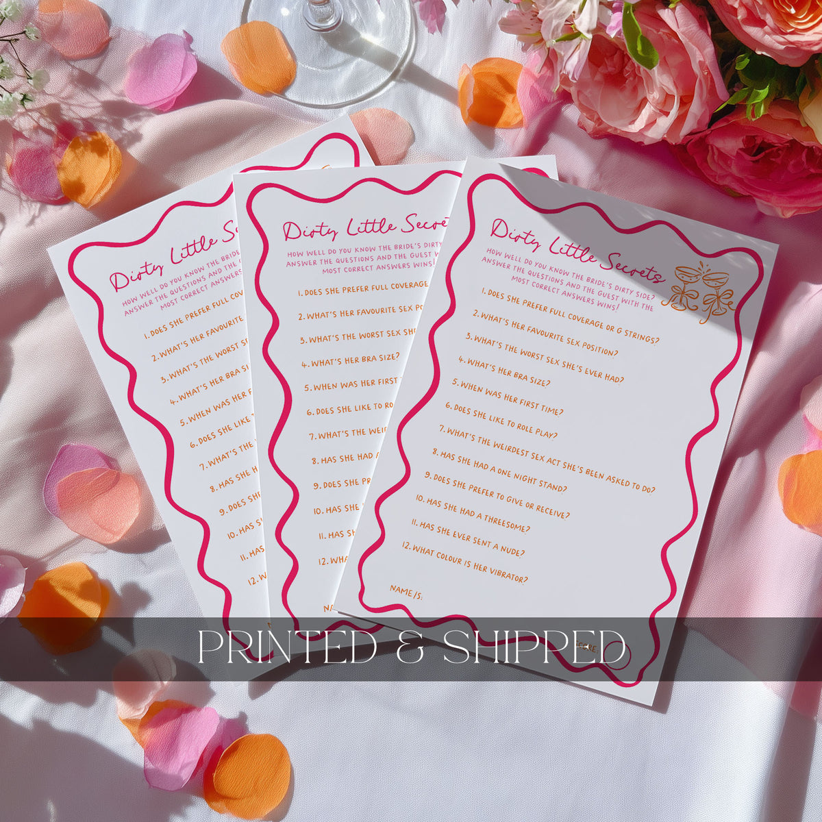 Three printed 'Scavenger Hunt' party game cards fanned out on a white surface with pink and orange flower petals, roses, and a wine glass in the background. A translucent banner at the bottom reads 'PRINTED & SHIPPED'.