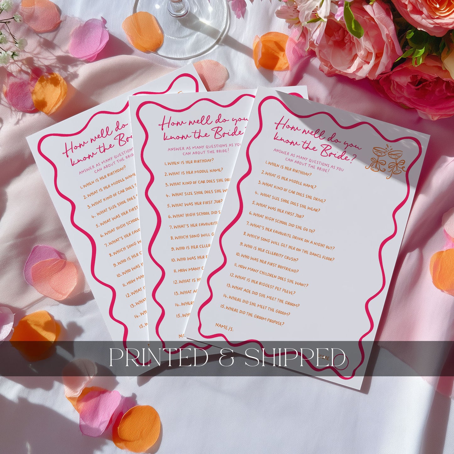 Three printed hens party game cards fanned out on a white surface with pink rose petals and flowers around them. The cards are white with pink wavy borders and gold text reading wedding-themed questions, with a 'PRINTED & SHIPPED' banner across the bottom.