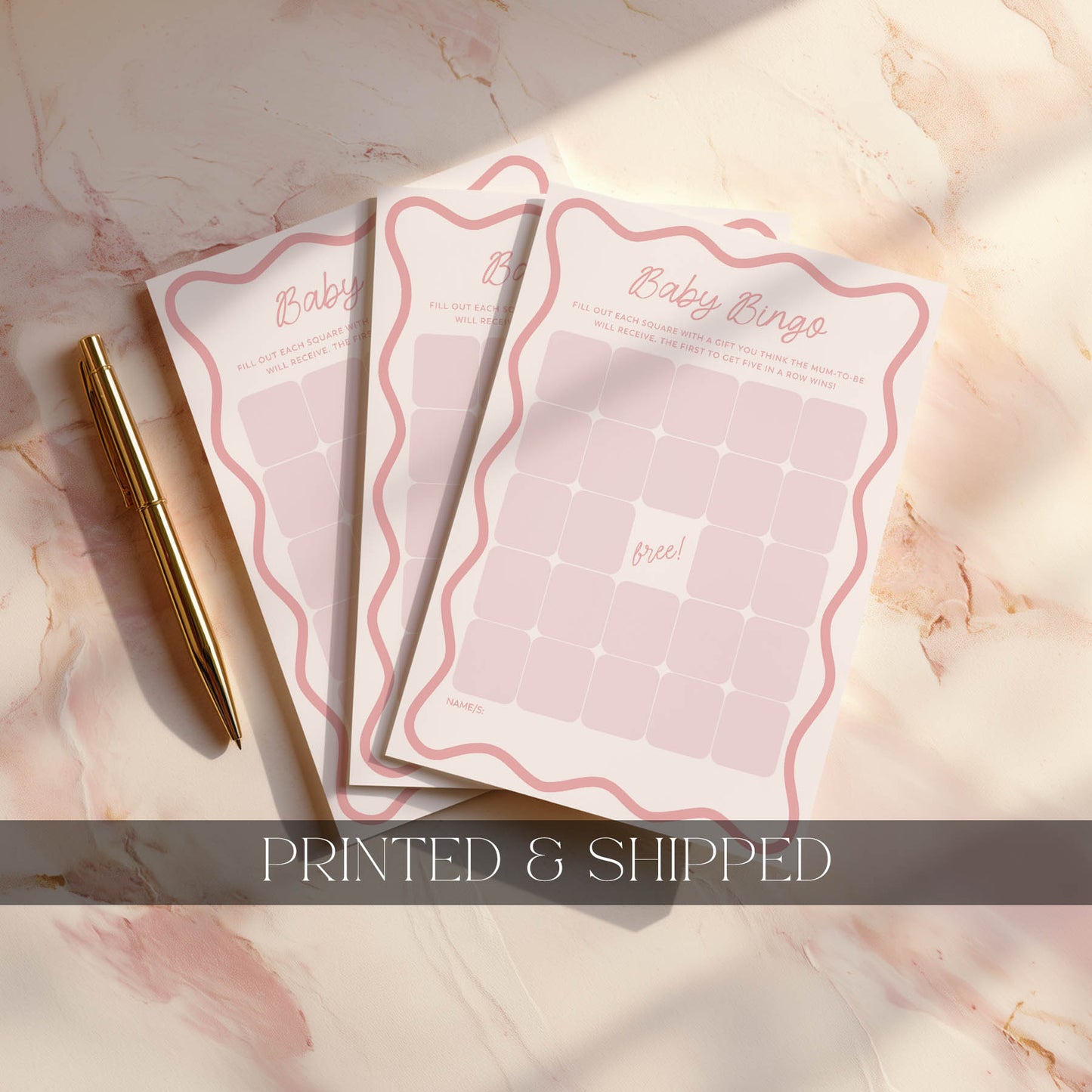 Three pink wavy baby bingo cards on a marble surface with a gold pen.
