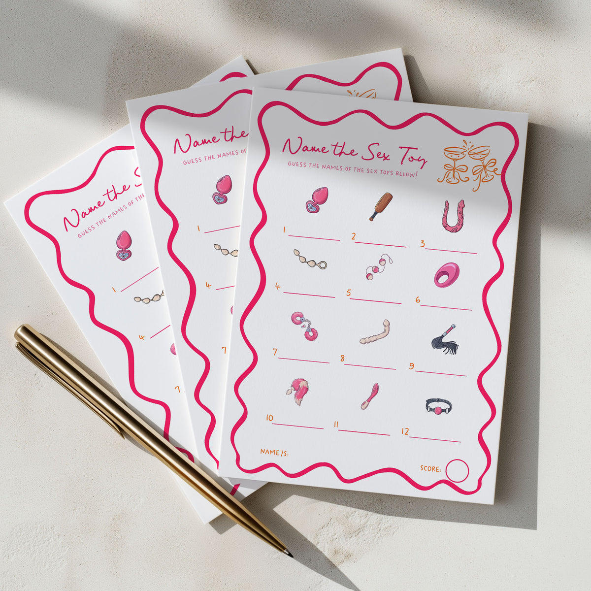 Three printed party game cards titled "Name the Sex Toy" are fanned out on a light surface, with a gold pen resting diagonally across the lower left. The cards are white with bright pink wavy borders and pink illustrated toy icons, photographed in soft natural light with shadows.