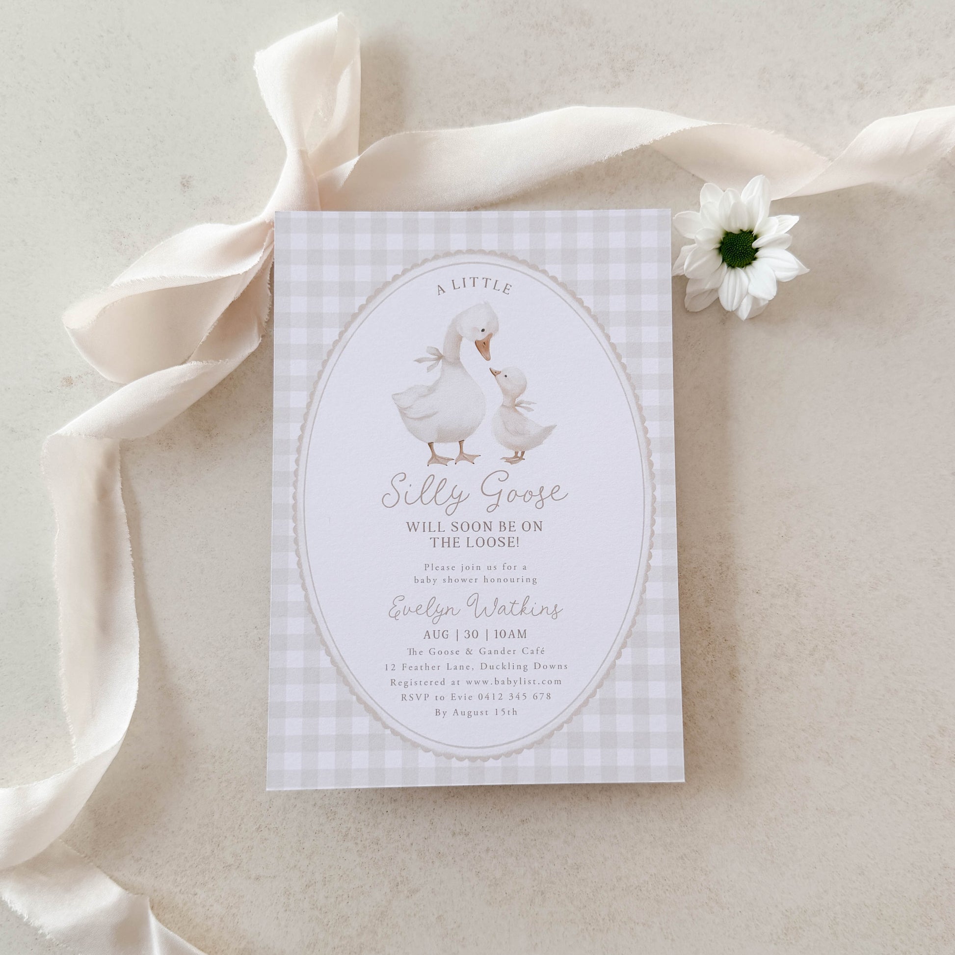 Silly Goose Baby shower invitation card with geese on a beige gingham background, placed on a light fabric surface.
