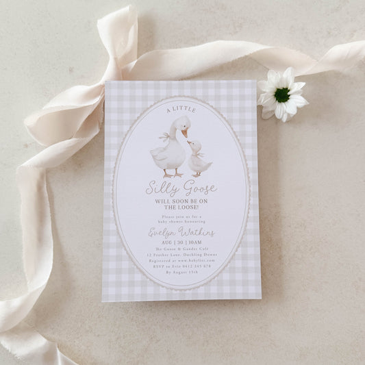 Silly Goose Baby shower invitation card with geese on a beige gingham background, placed on a light fabric surface.