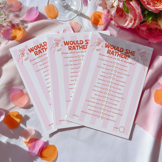 Three printed game cards titled "WOULD SHE RATHER" arranged on a white surface with pink and orange flower petals and flowers around them. The cards are pale pink with orange text and small line drawings, including a cocktail glass and a woman silhouette.