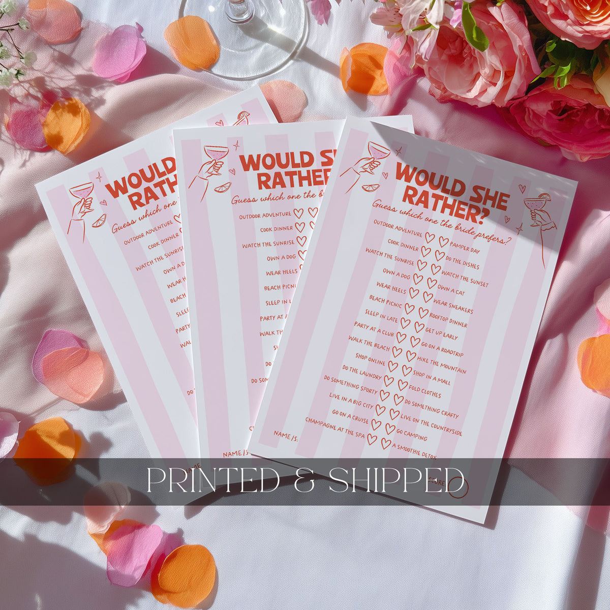 Three pink and white 'Would She Rather?' party games cards are displayed in a flat-lay arrangement with rose petals, flowers, and a wine glass on a white tablecloth. A dark gray banner at the bottom reads 'PRINTED & SHIPPED'.