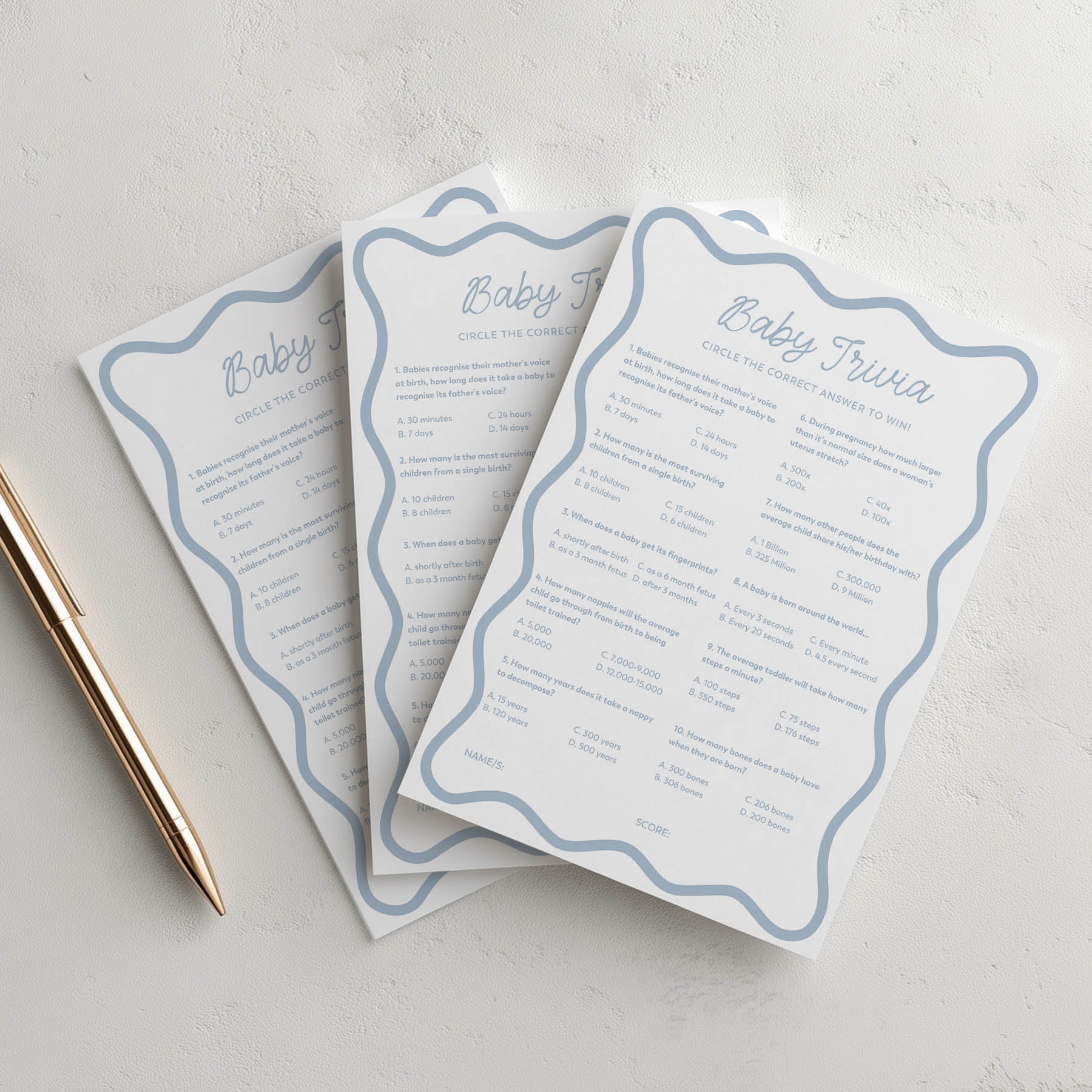 Three blue wavy sheets of baby trivia cards with a gold pen on a soft pink background